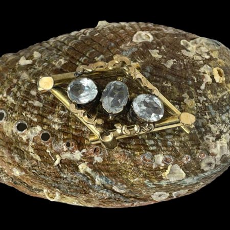 Main photo of Victorian Pinchbeck topaz brooch seen displayed on top of a shell with the brooch front forward facing. The main frame of the brooch is an elongated diamond shape with a vertical oval cut topaz stone to the centre with horizontally set stones either side. The stones are a beautiful light blue shade.