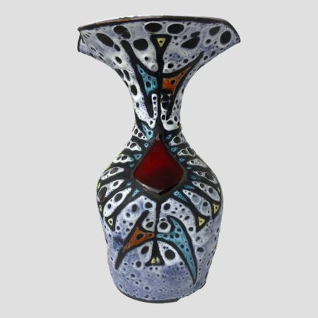 Main photo of Vallauris fat lava vase seen from an eye level with the cherry red glaze feature in the centre foreground. The main colour of the vase is a pale blue with yellow, orange and darker blue shapes with black piping. There are raised off white areas with black flecks. The vase shape is like a bottle on the lower half with a wide lip at top which has 2 spouts.