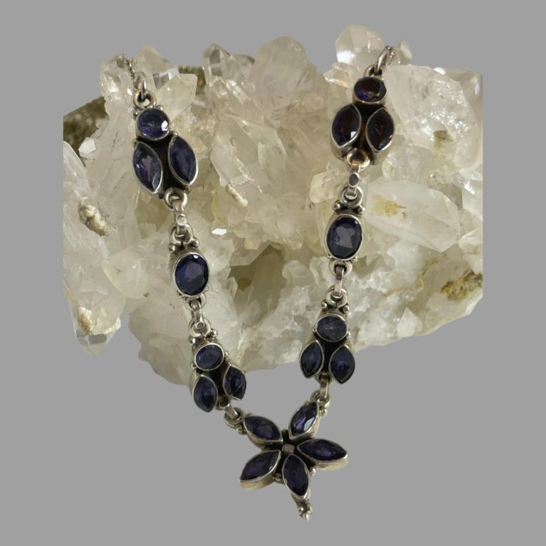 Main photo of sterling silver iolite necklace displayed laid across a rock of natural quartz crystal with just the front section housing the iolite stones on display. The central area of the necklace is a floral shape with 5 marquise cut iolite stones as petals. On either side of this flower is what could be a bee form-a round cut iolite with 2 vertically set marquise cut stones which could be wings. Next up is an oval cut stone followed by another bee like form. The iolite stones are slightly colour changing from a deep blue to a deep purple.