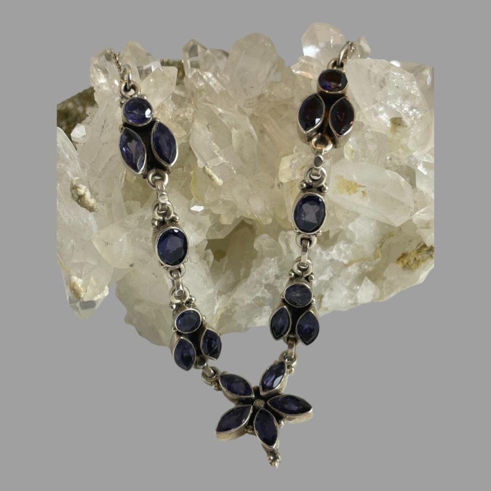 Main photo of sterling silver iolite necklace displayed laid across a rock of natural quartz crystal with just the front section housing the iolite stones on display. The central area of the necklace is a floral shape with 5 marquise cut iolite stones as petals. On either side of this flower is what could be a bee form-a round cut iolite with 2 vertically set marquise cut stones which could be wings. Next up is an oval cut stone followed by another bee like form. The iolite stones are slightly colour changing from a deep blue to a deep purple.