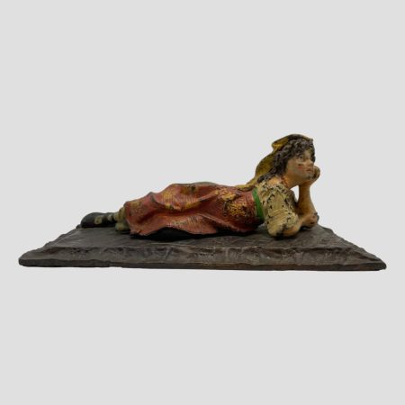 Main photo of vintage bronze Bedouin girl seen from the front side of her reclining position. She is lying on a striped rug shaped base of one plain dark grey colour with head resting on one raised arm and the other arm crossed in front of her. She has a dreamy look on her face. There are missing flakes of paint throughout which makes it look as though she has been playing in the dirt - this does add extra charm to this lovely piece.