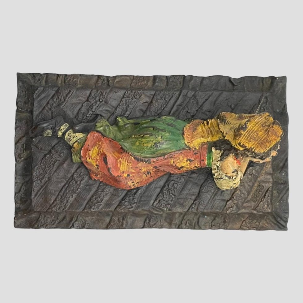 Photo looking down at vintage bronze Bedouin girl from above. Her feet are to the left and head to right and the shape and striped pattern of the 'rug' she is lying on is shown.