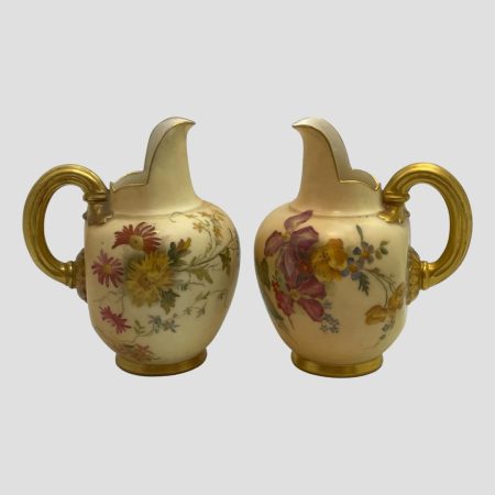 Main photo of Royal Worcester blush jugs shown side by side with the spout ends at the centre. The spouts are high as opposed to sideways of regular jugs. The sides with most flowers on are visible in the photo; to the left, daisy like flowers in yellow, pink and red and on the right clematis like flowers in purple pink and yellow with blue pansies and forget me nots. The handles, base and around the rim of the jugs is in gilt with some gilt highlights added to the floral images. For jug collectors and flower lovers alike - a beautiful pair of blush ivory jugs.