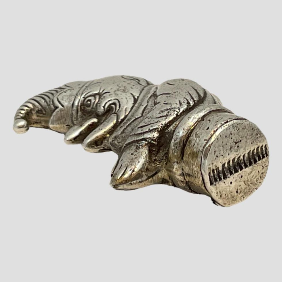 Photo of novelty silver Punch vesta shown laid flat on its side with the strike on the bottom displayed in the foreground. The vesta is laid diagonally across the page with the tip of Mr Punch's hat in the top left corner.