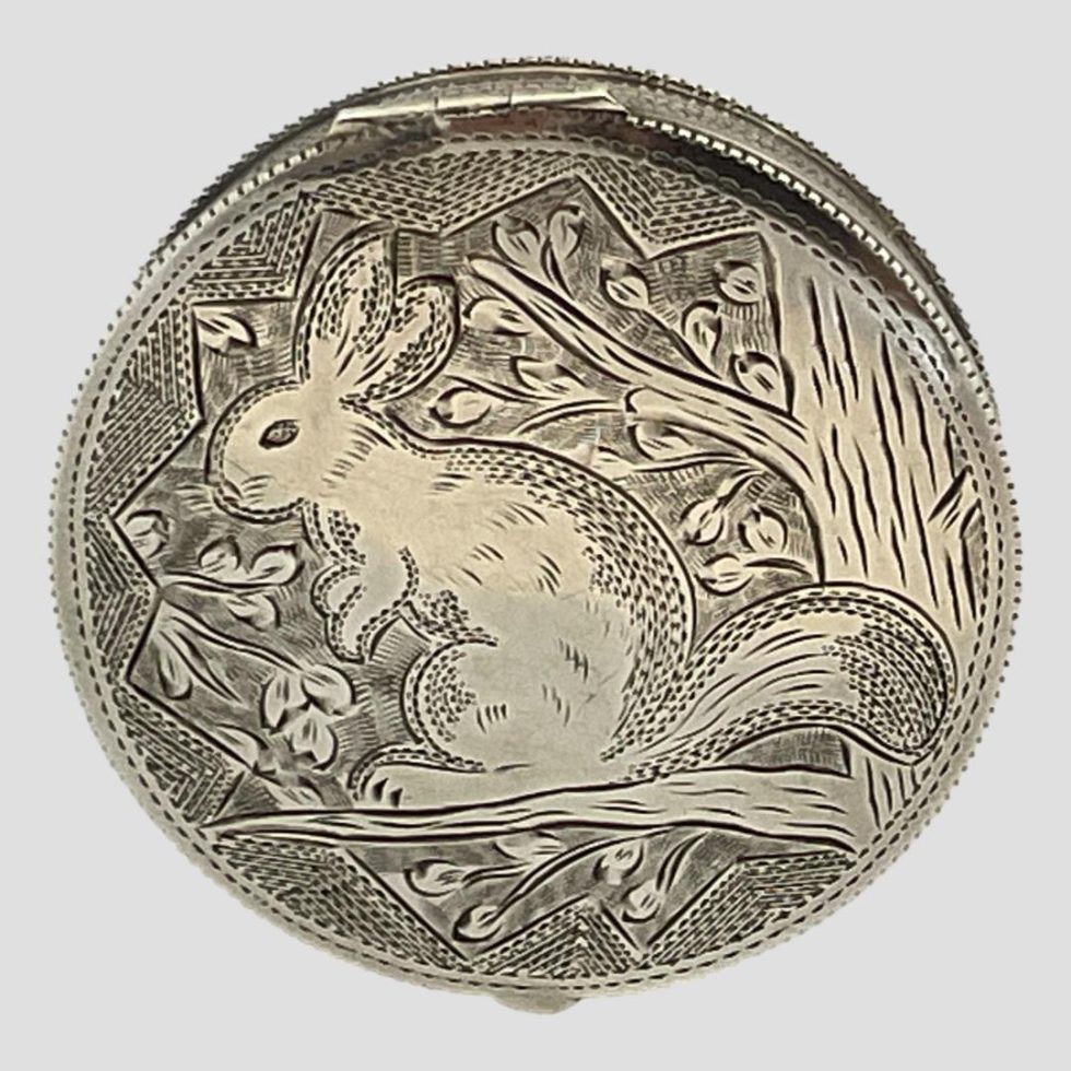 Photo of 1930s Austrian silver compact displayed upright on its side with the image of squirrel on the top of lid on display.