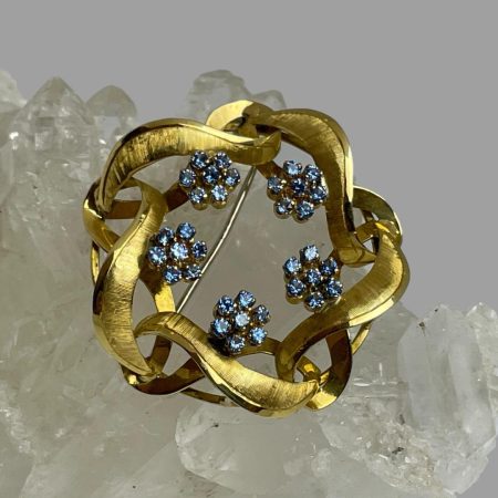 Main photo of gold blue topaz brooch displayed on top of a rock of natural quartz crystal with the brooch front forward facing. The gold is worked like a wreath with no centre and the 5 blue topaz flower heads are spaced out on the inside of the wreath line. The blue of the stones is unusually dark for topaz and look more like cornflower sapphire blue. The 18 karat gold and the topaz blue marry beautifully together.