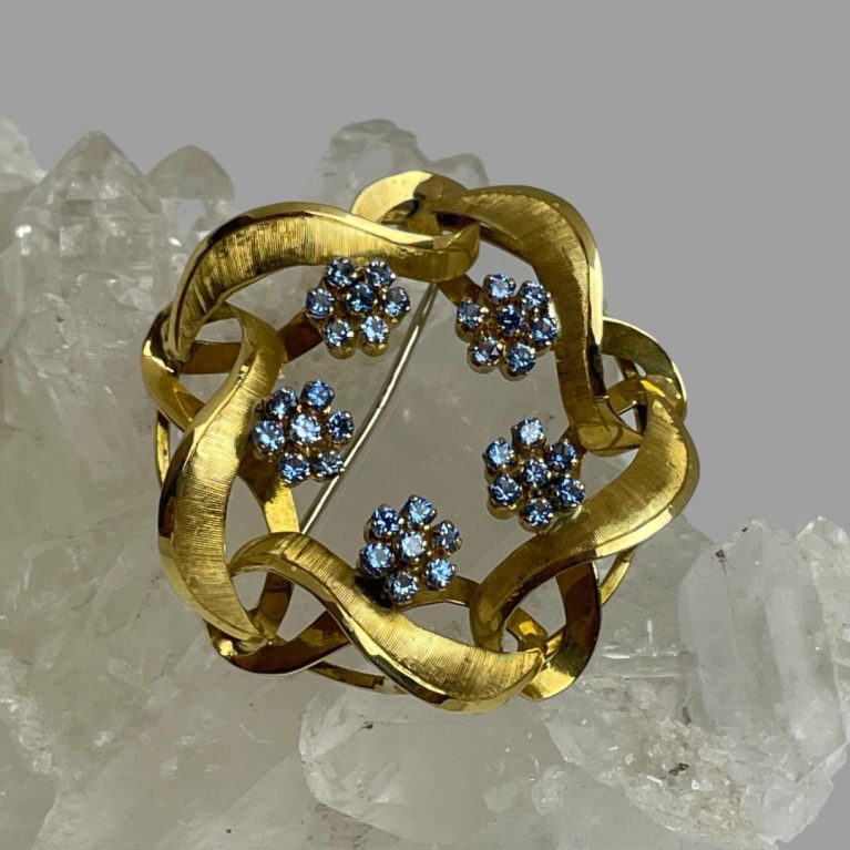 Main photo of gold blue topaz brooch displayed on top of a rock of natural quartz crystal with the brooch front forward facing. The gold is worked like a wreath with no centre and the 5 blue topaz flower heads are spaced out on the inside of the wreath line. The blue of the stones is unusually dark for topaz and look more like cornflower sapphire blue. The 18 karat gold and the topaz blue marry beautifully together.