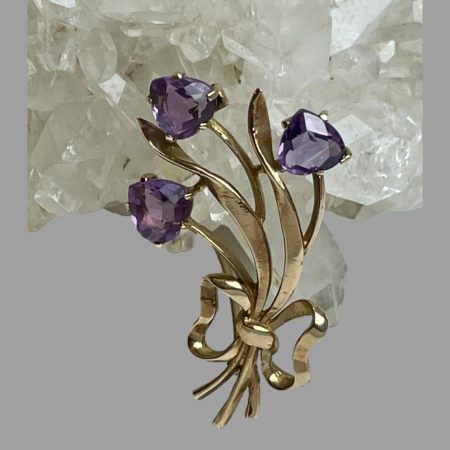 Main photo of gold amethyst tulips brooch shown leaning against a rock of natural quartz crystal with the 3 tulip flower heads at the top and gold ribbon holding the stems together at the base. The whole bunch leans slightly to the left as do their leaves. The amethyst are almost heart shaped but upside down hearts with pointed tip of tulip at the tops.