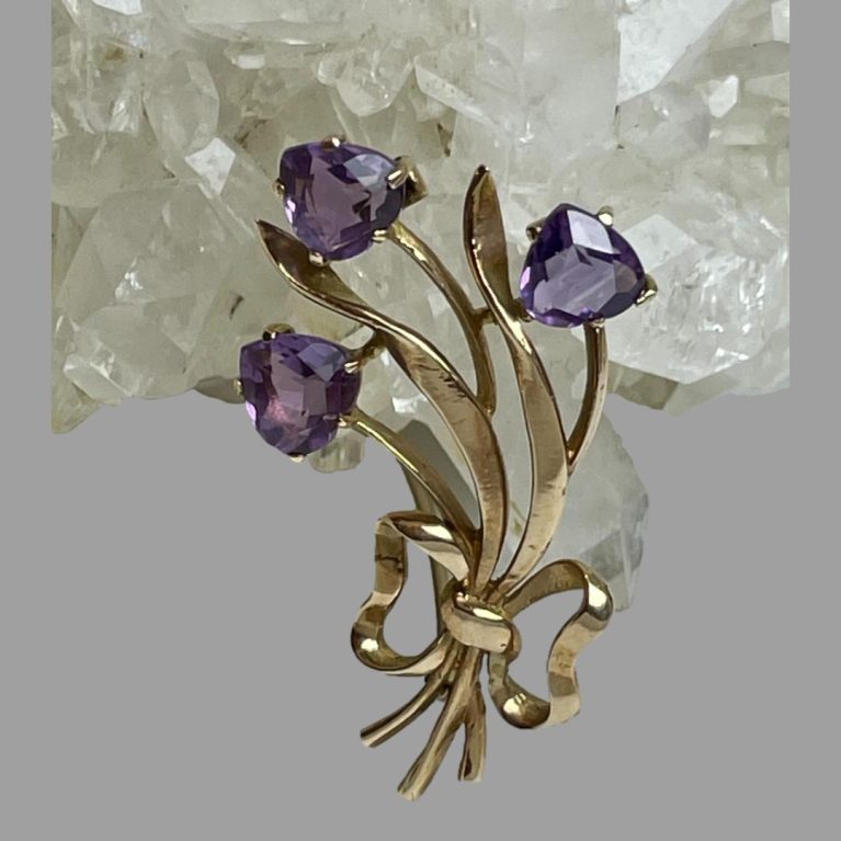 Main photo of gold amethyst tulips brooch shown leaning against a rock of natural quartz crystal with the 3 tulip flower heads at the top and gold ribbon holding the stems together at the base. The whole bunch leans slightly to the left as do their leaves. The amethyst are almost heart shaped but upside down hearts with pointed tip of tulip at the tops.