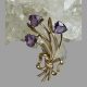 Main photo of gold amethyst tulips brooch shown leaning against a rock of natural quartz crystal with the 3 tulip flower heads at the top and gold ribbon holding the stems together at the base. The whole bunch leans slightly to the left as do their leaves. The amethyst are almost heart shaped but upside down hearts with pointed tip of tulip at the tops.
