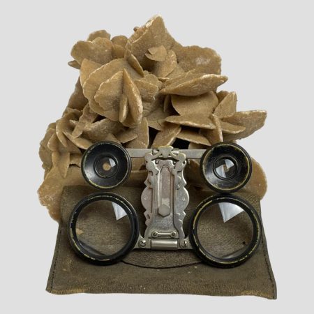 Main photo of French folding opera glasses shown displayed outside and on top of its leather case. The glasses are shown fully folded and therefore flat and they are leaning up against a rock of natural desert sand rose.