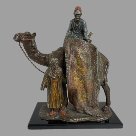 Main photo of spelter rug merchants (striker) showing the whole figurine from side where one merchant atop the camel has a rug rolled out on display whilst other merchant stands by the camels front leg holding the lower half of rug out in full 'look at our beautiful rug' selling pose. The camels head and neck are out to the left. The whole scene stands on a spelter base which is then on top of a black marble base. There is a fair bit of wear to the paint but the original colours still shine through in several places.
