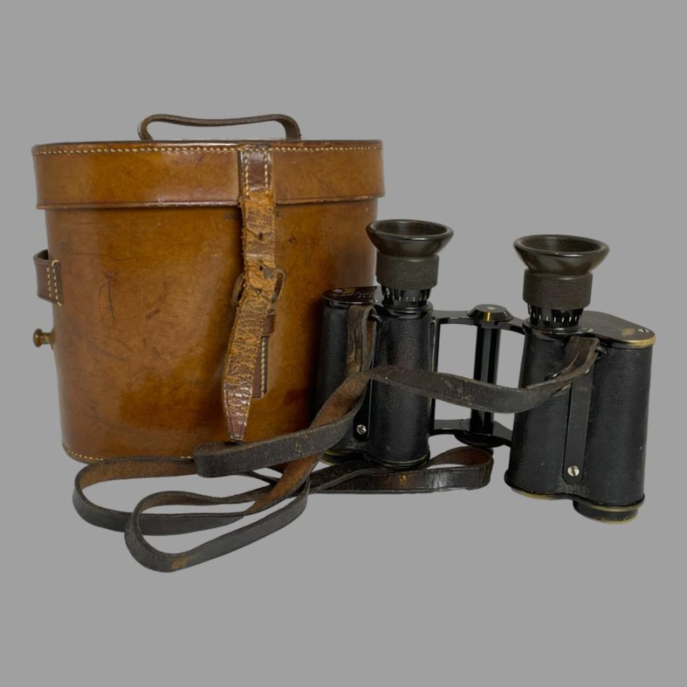 Photo of Carl Zeiss Jena binoculars displayed outside of their case. The tan brown leather case is on the left with the binoculars on the right and slightly to foreground. The binoculars are displayed lens down with the strap to the foreground and left.