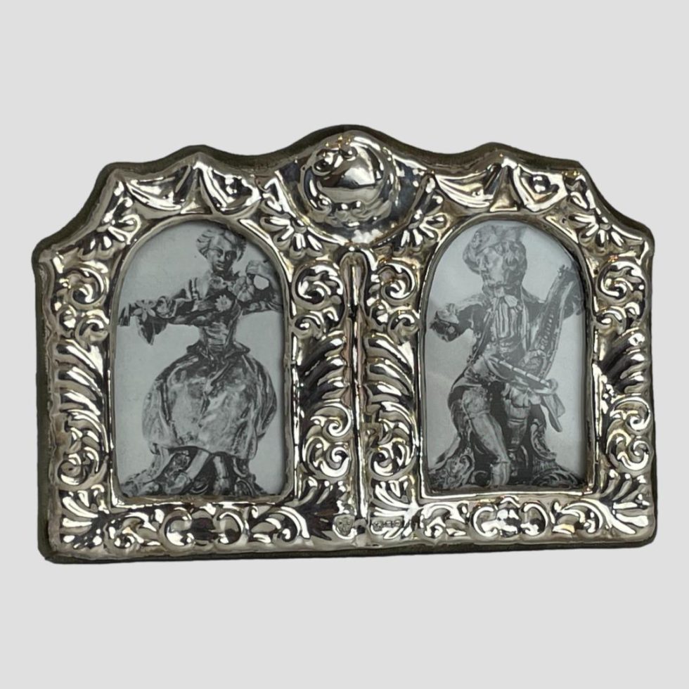 Main photo of small double photo frame seen forward facing. The sterling silver frame around the front of the frame is elaborately decorated with embossed detail. The area for the photos are small with an arched top.