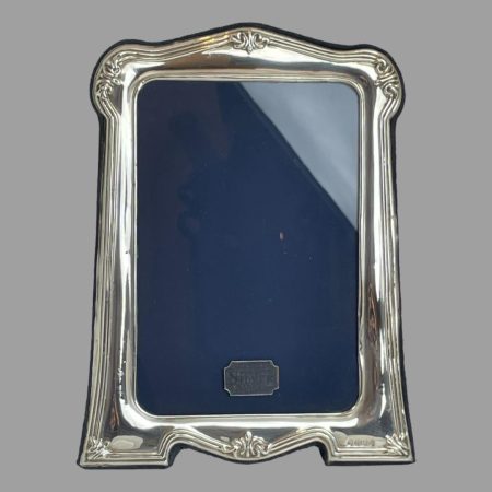 Main photo of Nouveau style photo frame seen with the front forward facing. The decorative detail on the silver is quite minimalist by Art Nouveau style standards but unmistakeably visible in a subtle capacity. The lower end of the frame is widest tapering a little at the top. The photo area remains a uniform rectangular shape. The minimal decoration consists of 3 raised lines with small ribbony areas to each corner as well as at the centre along the top and bottom. Definitely a frame that is simplistically pretty but also will not detract from the image at all. Nice gift item - just add photo!