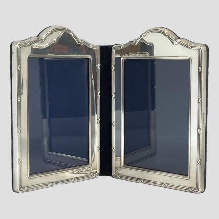 Main photo of silver double photo frame seen from an eye level with the 2 frames slightly inward facing to prop the frame open. The shape of the frames are a rectangular with a raised bump area to the top of each. The central folding area is blue velvet and this is the area with wear as it is most flexible. The design on the silver is what may be considered classical. 3 lines around the photo space area and 3 again on outer area except the outer lines have crosses spaced out all around. The area for the photographs is of a generous size