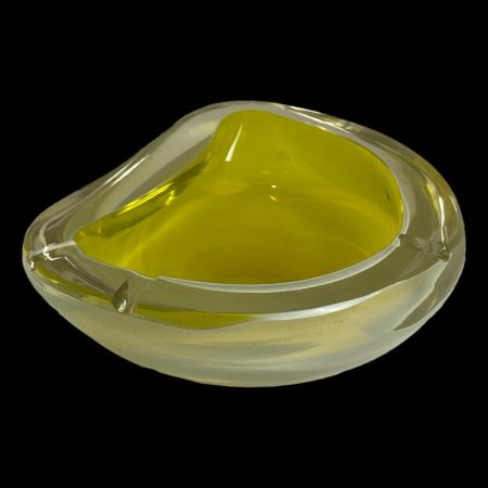 Main photo of Murano uranium glass ashtray seen from a slight side and raised angle. The flat side of the bowl is in the top right corner and the bulbous side in the bottom right. This ashtray has 2 cigarette rests on one side and 1 rest to the centre of bulbous area. Quite a mystery! The outside of ashtray is a milky opalescent colour whilst the inside is a lemon curd yellow. Set against a black background, these colours become more prominent.