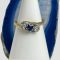 Main photo of 18kt sapphire diamond ring displayed on a cone shaped stand with the ring front forward facing. The stones are set in a diagonal formation along the split band which gives an impression of 2 arms holding the gems. A small slice of blue agate is behind the ring.