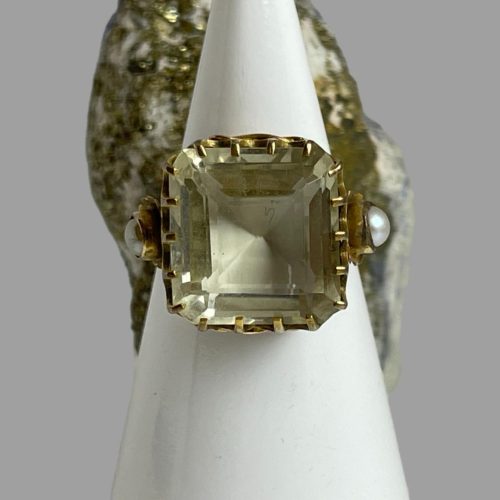 Main photo of gold citrine dress ring displayed on a white cone shaped stand with the front of ring forward facing. The citrine is a lovely pale minty yellow colour and the step cut of the stone adds extra quality to the ring. There are a total of 18 prongs holding the stone in place (4 above & below and 5 on left and right) There is a small rock of natural fools gold behind the cone stand. The split pearls to either shoulder are prominent.