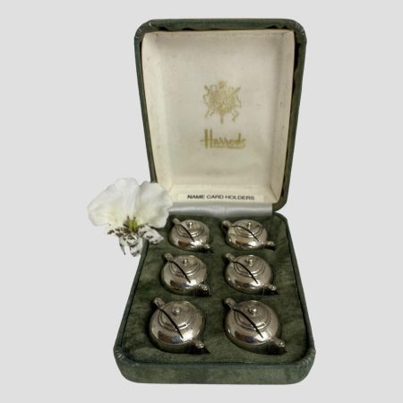 Main photo of Harrod's name card holders displayed inside their fitted green velvet case. The fitted slots hold each teapot at a diagonal setting so each teapot sits neatly in a diagonal row (3 to each side) The Harrod's crest and name are stamped on the cream silk interior of the lid. A single white orchid has been added to the photo to the left.