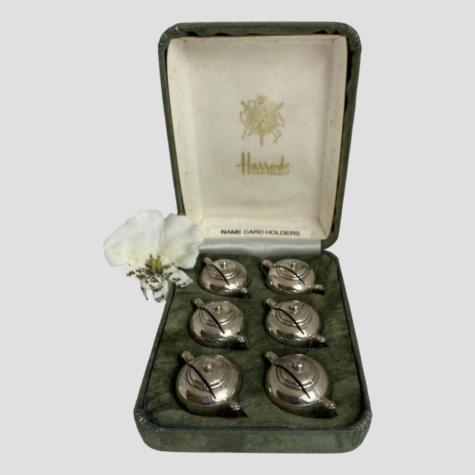 Main photo of Harrod's name card holders displayed inside their fitted green velvet case. The fitted slots hold each teapot at a diagonal setting so each teapot sits neatly in a diagonal row (3 to each side) The Harrod's crest and name are stamped on the cream silk interior of the lid. A single white orchid has been added to the photo to the left.