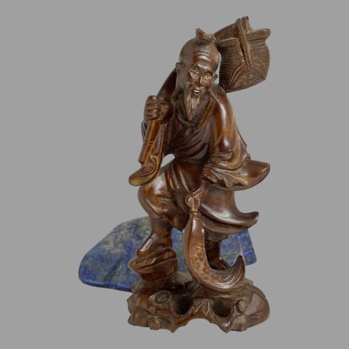 Main photo of carved wooden Oriental fisherman seen from the front. The fishermans body is facing the camera but his head is slightly to the right. He is carrying a large carp which is being carried in his left hand and his right hand is holding his fishing rod which is held over his shoulder with a basket on the end. There is a serene happy look about his face. The boxwood is a warm chestnut brown colour and the carving detail is of good quality. There is a rock of blue lapis lazuli in the background to offer a feeling of a lake.