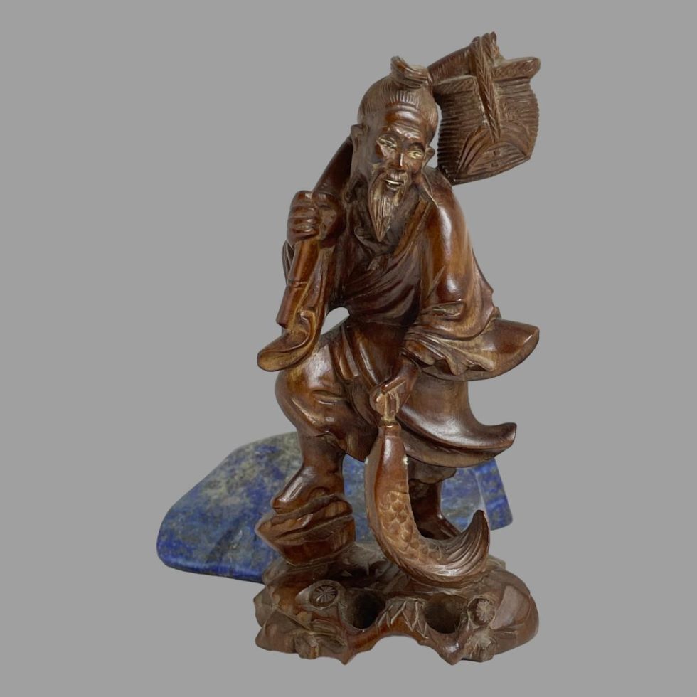 Main photo of carved wooden Oriental fisherman seen from the front. The fishermans body is facing the camera but his head is slightly to the right. He is carrying a large carp which is being carried in his left hand and his right hand is holding his fishing rod which is held over his shoulder with a basket on the end. There is a serene happy look about his face. The boxwood is a warm chestnut brown colour and the carving detail is of good quality. There is a rock of blue lapis lazuli in the background to offer a feeling of a lake.
