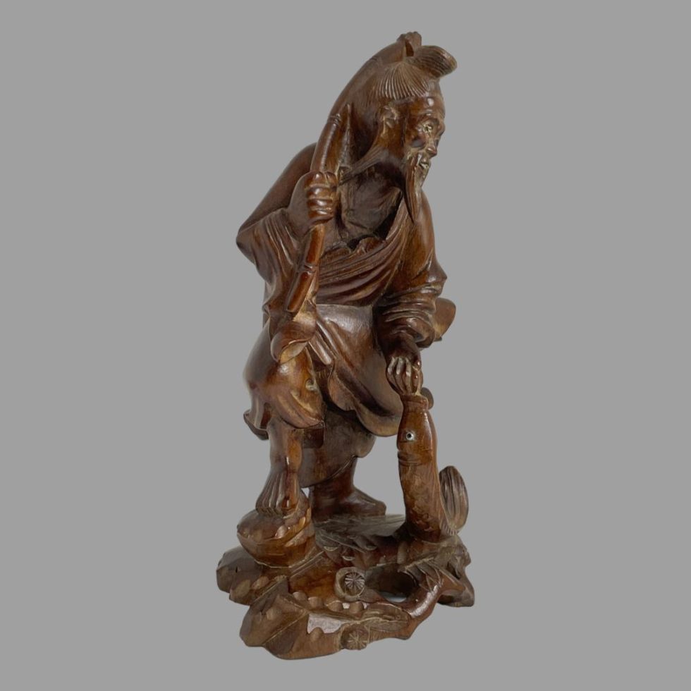 Photo of carved wooden Oriental fisherman seen from a side angle. His right foot is in the foreground and his head is faced towards the right. There appear to be carved wooden flower heads at his feet.