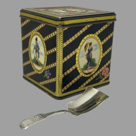 Main photo of Antique tea caddy spoon displayed laid flat in front of a tea caddy tin. The spoon has a shovel like shape and is typically short handled. The tin is for display purposes only.