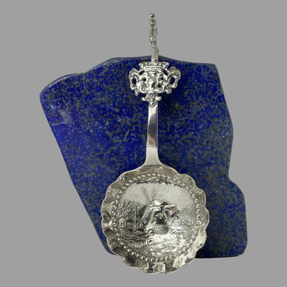 Main photo of Dutch silver ladle spoon leaning upright against a rock of natural lapis lazuli. The spoon bowl has a skirted rim which is separated from the centre of bowl with silver 'jewel' (dots) The windmill takes pride of place in the bowl centre with other buildings in the background which include a church. Just before the tip of the handle there is am elaborate piece of pierced silver a little like a fob on a watch chain which acts as a stabiliser for the hook back. The handle tip has a figure of what appears to be a schoolboy with satchel although could also be a postman perhaps. A lovely item for any silver collection and a lovely gift piece.