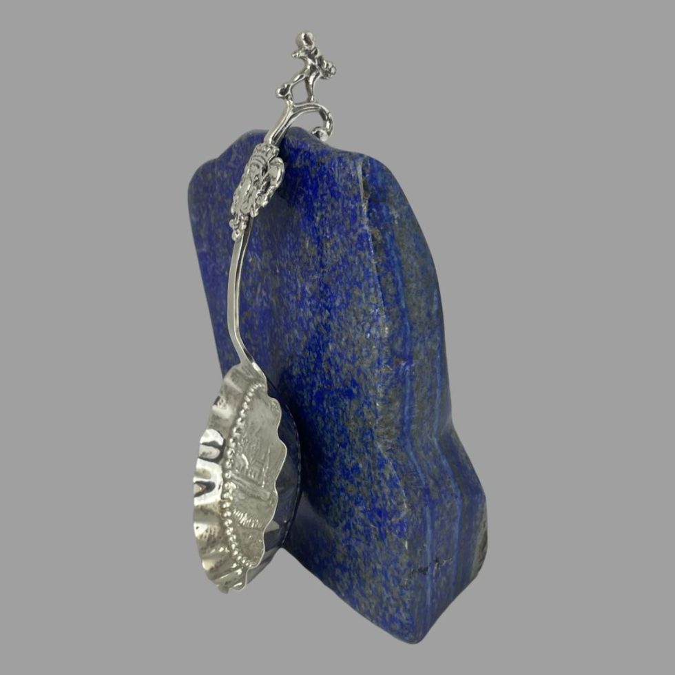 Photo of the Dutch silver ladle spoon still leaning against the rock of lapis and this time shown from a diagonally sideways angle so the spoon bowl is facing bottom left corner. The curve of the spoon is clearly visible.