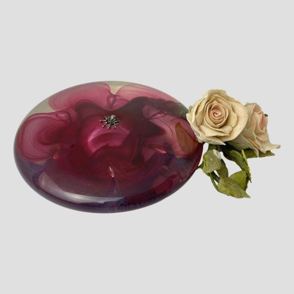 Main photo of pink lotus art glass seen from a raised angle looking diagonally down at the top. The piece is a little like a squished paperweight. It is circular and domed with a vibrant fuchsia pink lotus flower inside clear glass. The spider on the top is miniscule. A small posy of pale pink roses are laid to the right for effect.