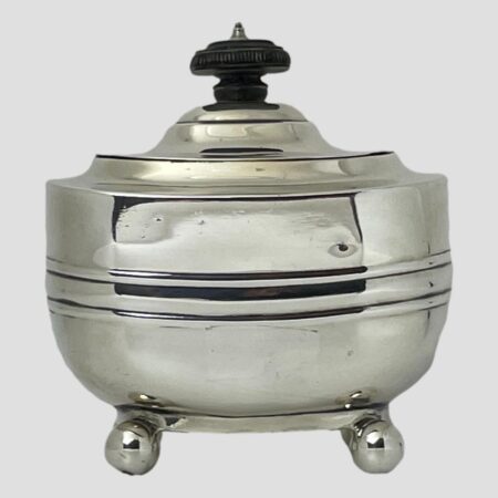 Main photo of Edwardian silver tea caddy seen from the front from an eye level. The caddy has a nice boat like shape to its lower part with round ball feet (2 visible) The bakelite finial has a drawer pull quality to it making it a breeze to open the lid which is on hinges.