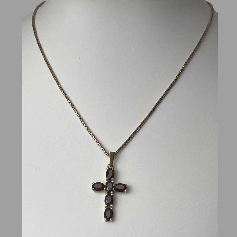 Main photo of gold garnet cross & chain displayed on a stand and seen with front of cross facing forward. The chain is a box chain of good thickness. The pendant has a smooth bail with 4 oval cut garnets going down and an oval cut garnet horizontally for the arms on the left and right so total of 6 garnets. Each garnet is held in place with 4 prongs so a nice secure setting.