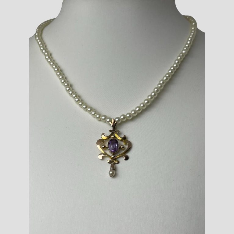 Main photo of Edwardian gold pendant collar seen displayed on a necklace display stand. The collar is forward facing with the pendant on full display. The pendant has a outer gold frame with a fleur-de-lis bail at the top and a beautiful oval cut amethyst to the centre. A single pearl hangs below finishing off the dreamy princess look. Thew collar consists solely of faux pearls with a small amethyst finishing off at either end. Very dreamy Princess look about this lovely necklace.