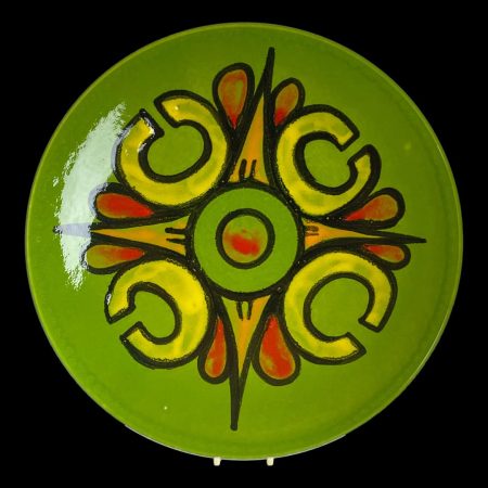 Main photo of large Poole Delphis dish displayed on a plate stand against a black background. The base colour for this dish is a nice leafy green - not dark. To the centre are colours of red, orange and yellow with pop art like shapes; bold letter C in yellow, bold green O in the centre and pointed orange arrow head like shapes with red seeds to their sides. A lovely smile inducing piece!