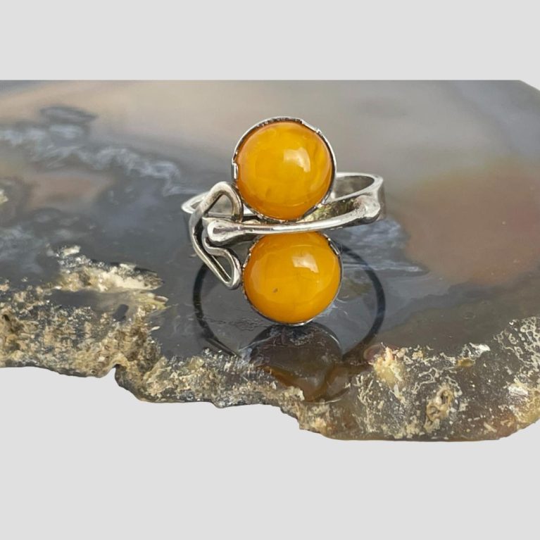 Main photo of vintage silver amber ring shown displayed on a slice on agate with the ring front forward facing. The egg yolk amber pieces are set like a figure of eight one on top of other and either side of the silver band. Across the silver band is an overlaid silver piece of a short bar with small ball ends then on one side of this bar is a heart like shape forged from another piece of silver. The band is flat with no fancy detail to shoulders.