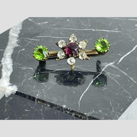 Main photo of vintage Suffragettes inspired brooch shown displayed on top of a slab of black marble. The brooch front is forward facing. At the centre is a flower with 6 'petals' of clear glass with purple glass centre and bright green glass at either end. What makes this brooch stand out is that all the glass pieces are round cut which somehow adds extra charm.