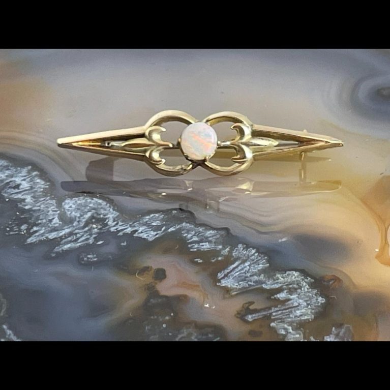 Main photo of antique gold opal brooch displayed on top of a slice of natural agate with the brooch front facing forward. The brooch has pointed ends like elongated Vs with rounded ends formed in the middle where the opal is housed. Within the rounded gold ends is a fleur-de-lis giving impression this piece is possibly of French origin.