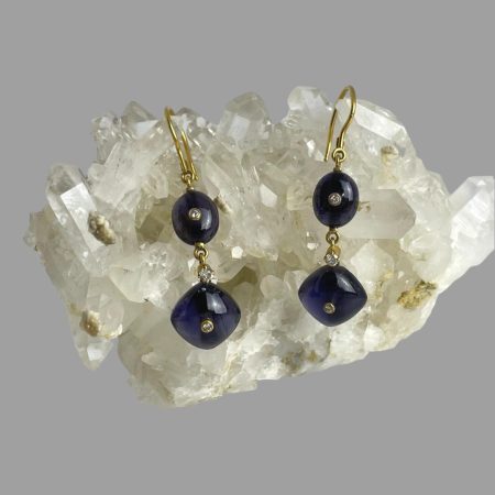 Main photo of gold iolite diamond earrings shown displayed hanging on a rock of natural quartz crystal with the earring fronts facing forward. The earrings construction begins with an 18 karat gold hook fastener. Am oval iolite cabochon with a small round cut diamond to the centre is first followed below it by a larger round cut diamond divider. At the bottom of the earring is a square iolite cabochon set with one corner at top like diamond shape and there is another small round cut diamond to the centre of this. The iolite is a beautiful darker blue with tinges of purple in certain light.