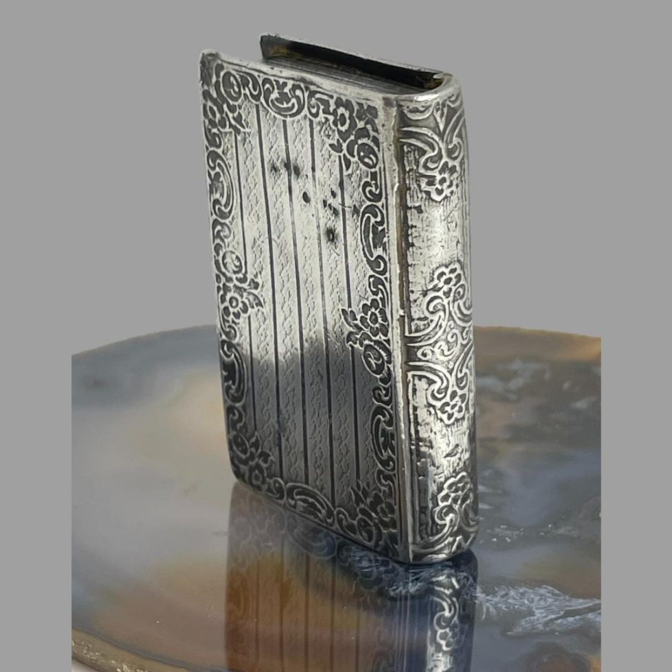 Photo of book shaped snuff box displayed on top of a slice of natural agate with the binding edge facing to the bottom right corner of photo and 'back of book' visible on left side.