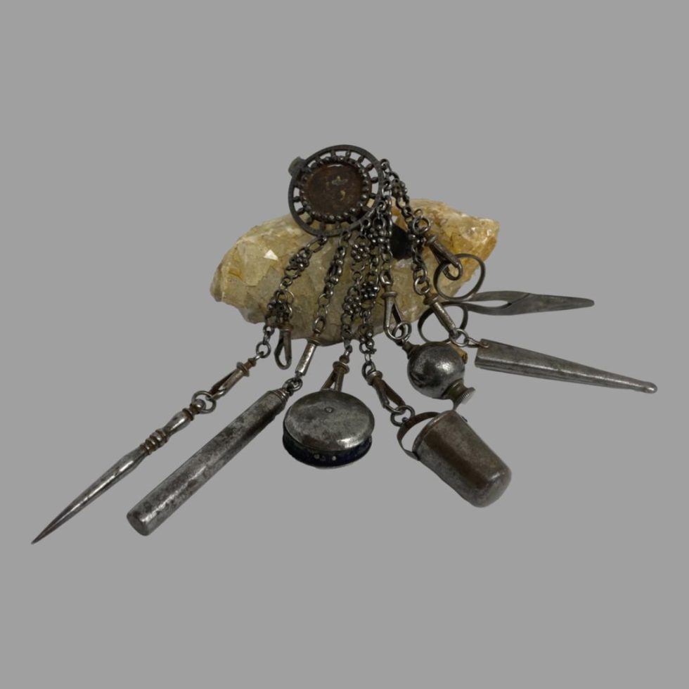 Main photo of antique steel chatelaine displayed strewn over a rock of natural river crystal. The top part of the chatelaine where the hook area is located is on top of the crystal rock and the tools hanging from it are fanned out in the foreground. From left to right, piercing tool, pin cushion, thimble, tape measure, Scissors cover and scissors.