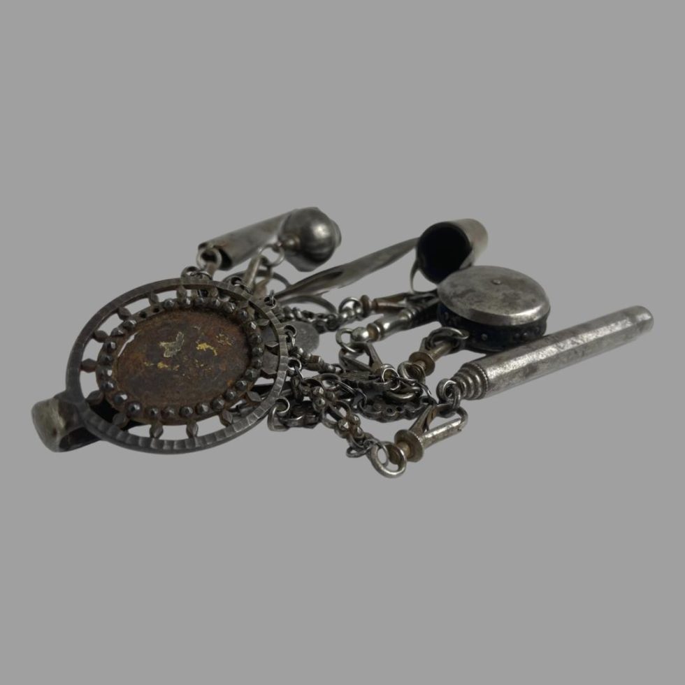 Photo of antique steel chatelaine displayed on a flat surface with the hook end in foreground to the left and the hanging tools laid towards the top right of photo. The front of the hook area is shaped as an oval and clearly missing a stone or perhaps even a cameo. It would elevate the quality of this piece to have something added there.