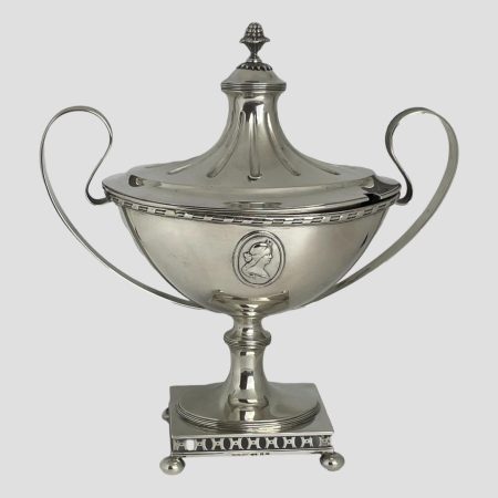 Main photo of Swedish silver soup tureen shown from a near eye level and from one side. Both sides are identical except for the lid which has a gap for the ladle (not included) This is an incredibly classical soup tureen which almost resembles first prize cup ! The base is a short rectangle with 4 ball feet, a short stem rises from its centre which holds a boat shaped tureen at its end. The cartouche of a lady is to the centre and the handles to left and right are delicate strips of silver moulded into an ear like shape. The lid fits nicely over the boat shaped tureen and has a tall centre not unlike a marquee top. This is definitely a talking point for any dining table and a wonderful piece for any silver collection.