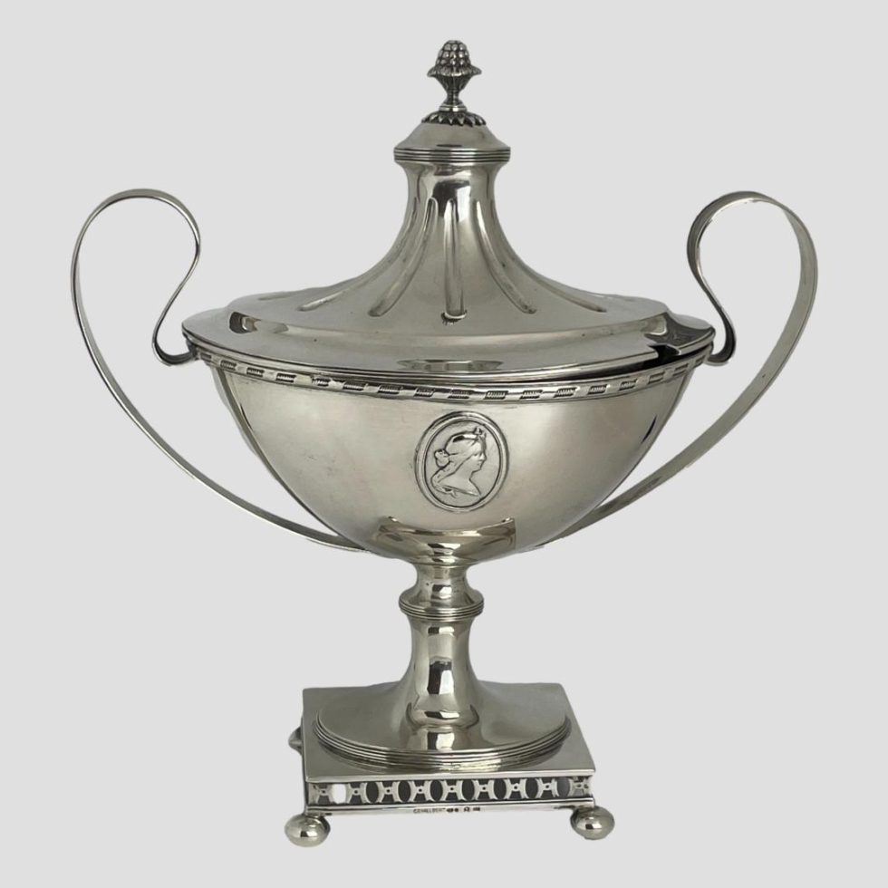 Main photo of Swedish silver soup tureen shown from a near eye level and from one side. Both sides are identical except for the lid which has a gap for the ladle (not included) This is an incredibly classical soup tureen which almost resembles first prize cup ! The base is a short rectangle with 4 ball feet, a short stem rises from its centre which holds a boat shaped tureen at its end. The cartouche of a lady is to the centre and the handles to left and right are delicate strips of silver moulded into an ear like shape. The lid fits nicely over the boat shaped tureen and has a tall centre not unlike a marquee top. This is definitely a talking point for any dining table and a wonderful piece for any silver collection.