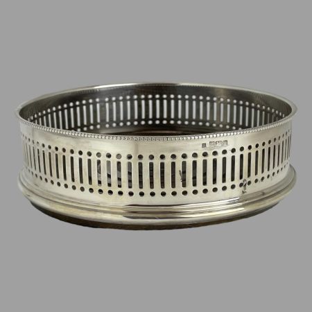 Main photo of sterling silver bottle coaster seen from an eye level and focusing on the silver gallery with pierced work all round. The full hallmark is visible just to the centre right around top rim of coaster.