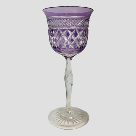 Main photo of rare amethyst crystal glass seen from an eye level. The glass is criss-cross cut at lower area of bowl with a second row of smaller criss-crosses. The base of the bowl has a daisy shaped design and the base has a sunburst cut. The amethyst colour is a dark lilac. Lovely as a gift with a bottle of wine.