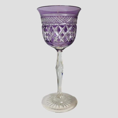 Main photo of rare amethyst crystal glass seen from an eye level. The glass is criss-cross cut at lower area of bowl with a second row of smaller criss-crosses. The base of the bowl has a daisy shaped design and the base has a sunburst cut. The amethyst colour is a dark lilac. Lovely as a gift with a bottle of wine.