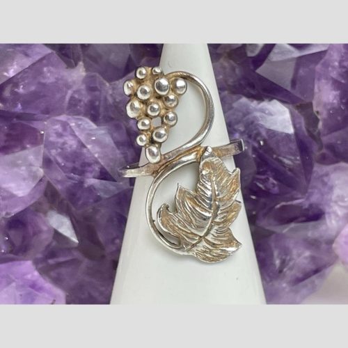 Main photo of ring displayed on a cone shaped stand with the ring front facing forward. The grape bunch is at the top hanging left while the vine curls over to the right and down like the number eight. The single vine leaf is at the end of the vine and opposite of where the grape bunch is. There is a rock of natural purple amethyst behind the ring.