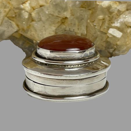 Main photo of silver & carnelian pill box seen from an eye level. The box is displayed in front of a rock of river quartz which is has a pale yellow crystal hue. This is reflecting on the silver area of the pill box lid. As with a lot of pill boxes with a stone set in the lid, this one has more depth to the lid than the base.
